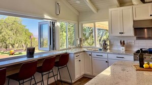 Una cocina con armarios blancos, encimera de mármol y vistas a árboles.