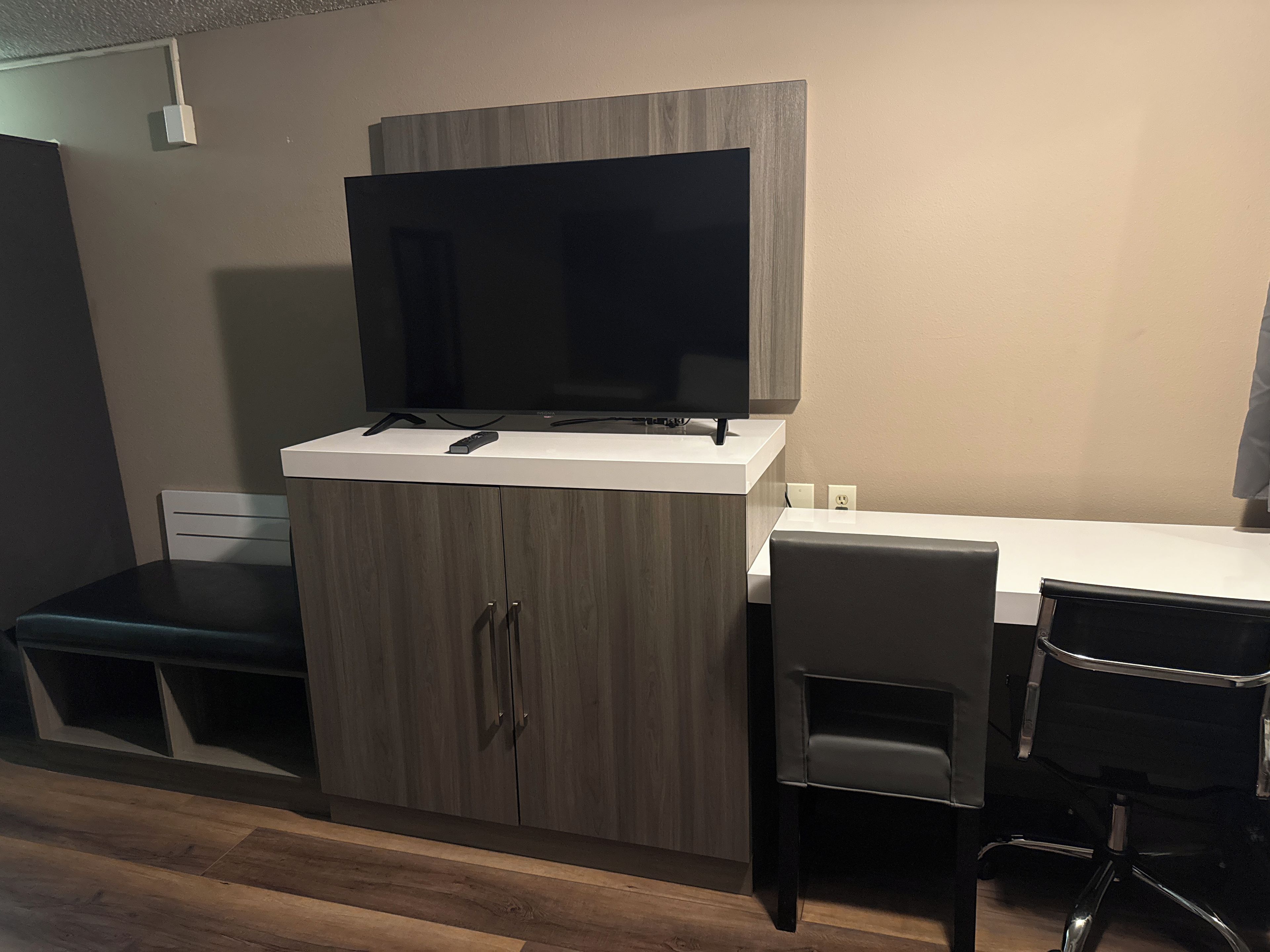 Een moderne kamer met een flatscreen televisie bevestigd aan een houten kast, een wit bureau en twee zwarte stoelen.