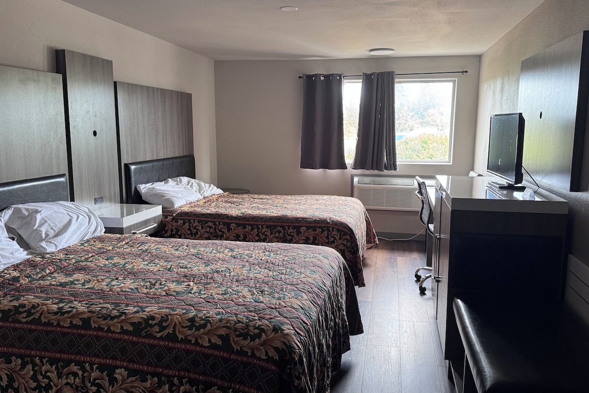 Ein Hotelzimmer mit zwei Betten, einem Schreibtisch, einem Fernseher und einem Fenster mit Vorhängen.