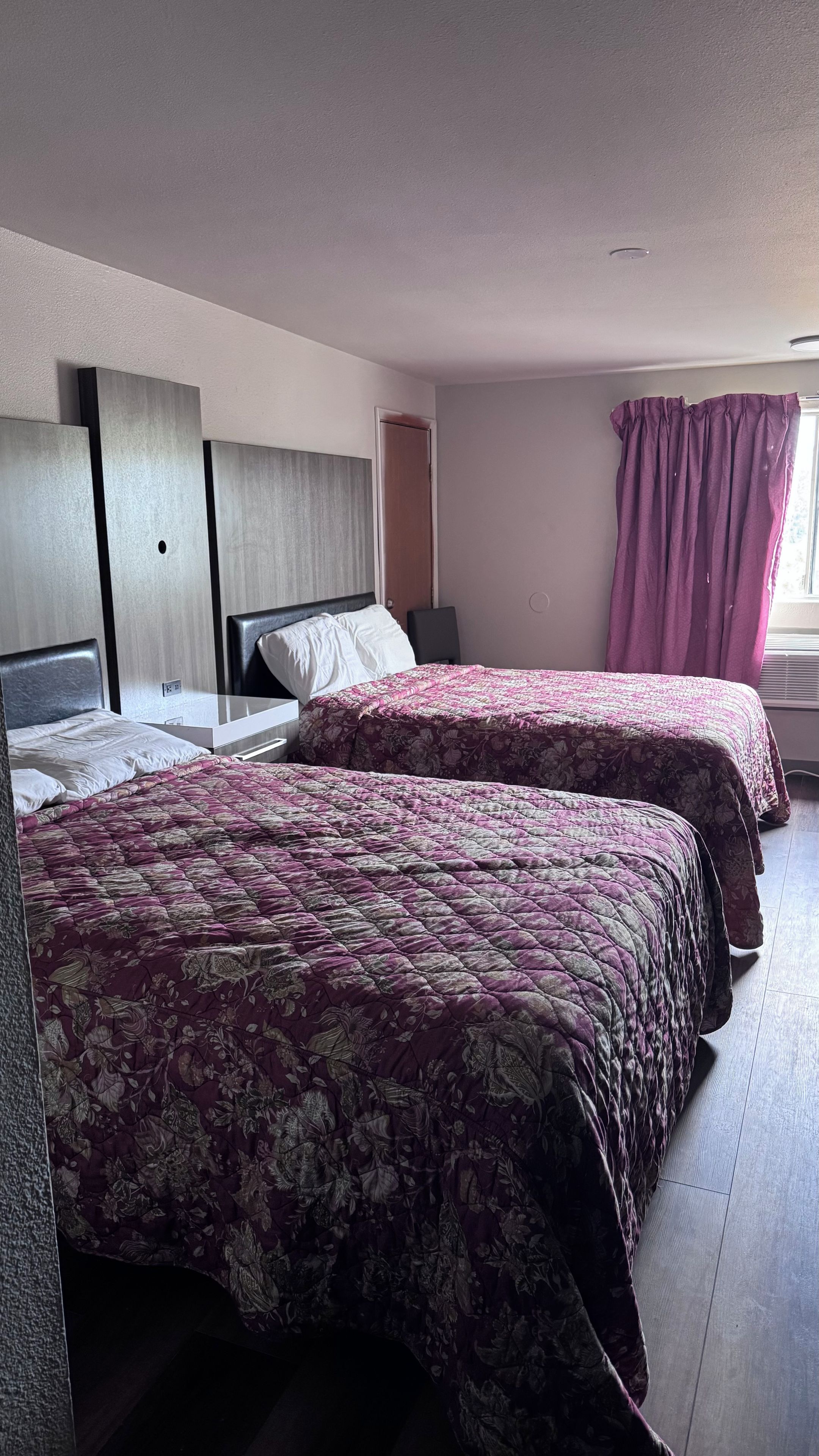A hotel room with two beds, a bedside table, a nightstand, a window with curtains, and a ceiling light.