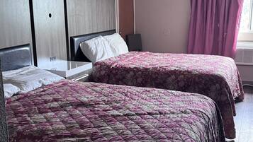 A hotel room with two beds, a bedside table, a nightstand, a window with curtains, and a ceiling light.