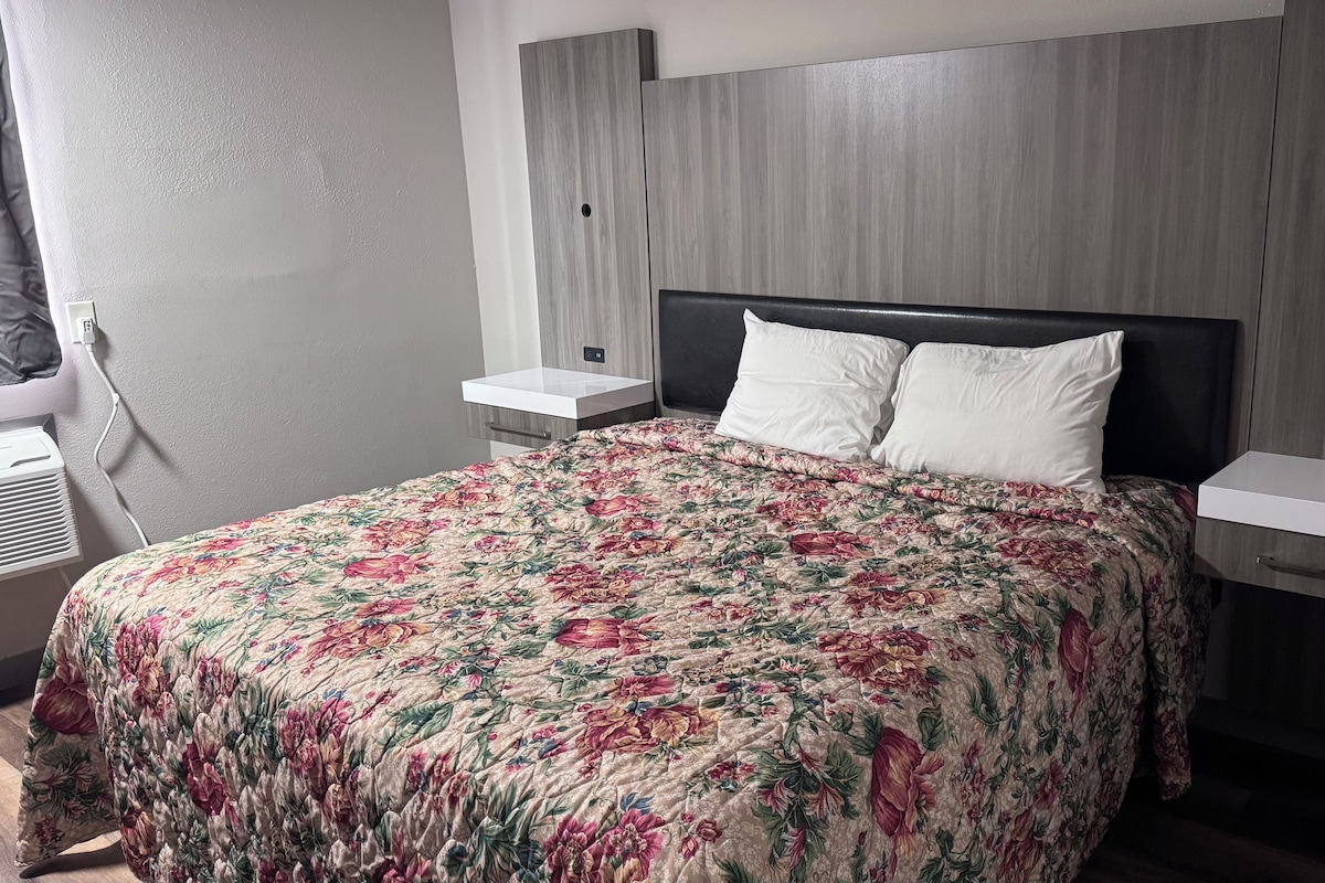 Ein Hotelzimmer mit einem Bett, Nachttischen, einem Fenster mit Vorhängen und einem an der Wand befestigten Regal.