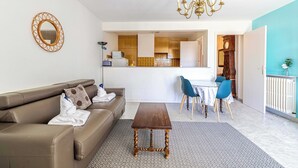 Una sala de estar con un sofá marrón, una mesa de centro de madera, un comedor con sillas azules y una cocina al fondo.