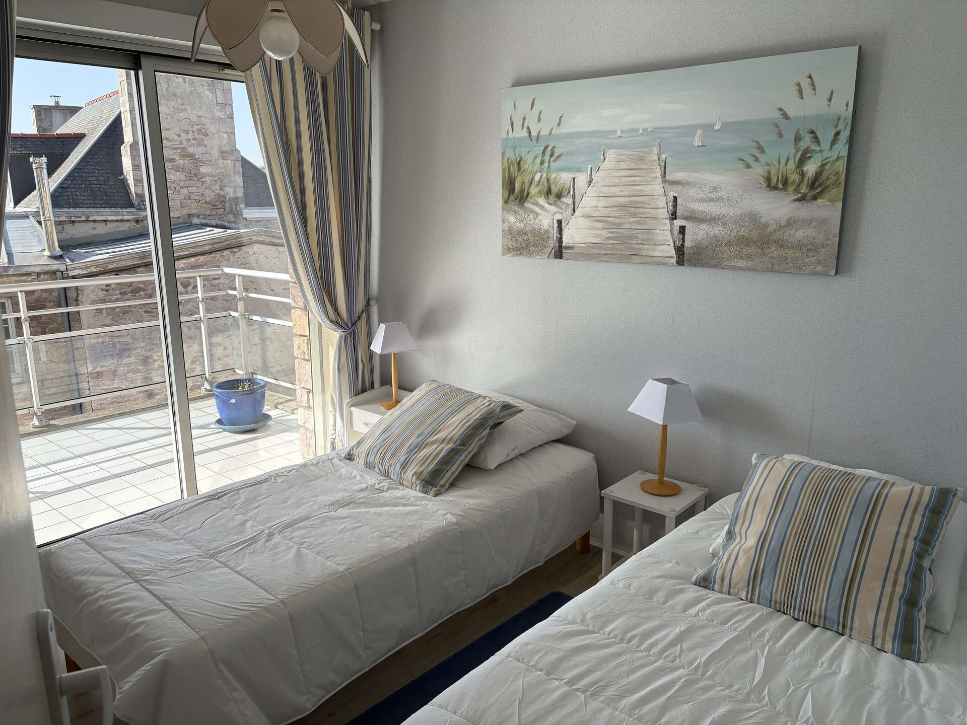 Ein Schlafzimmer mit zwei Betten, einem fenster mit Vorhängen, einem Gemälde mit Strandmotiv und Blick auf ein Gebäude.