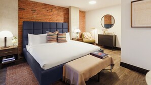 Une chambre d’hôtel avec un mur en briques, un grand lit avec une literie blanche, une table de chevet avec une lampe, une commode et un miroir au mur.