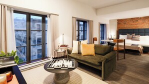 Une chambre d’hôtel moderne avec un canapé vert, une table basse ronde, un lit adossé à un mur en briques et de grandes fenêtres.