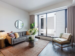 A modern living room with a grey sofa, a round coffee table, a side table, a chair, a large window with blinds, and a mirror on the wall. - Loft | Appartement paisible Vieux-Montréal, cour intérieure, cuisine équipé (Montréal)