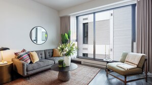 Een moderne woonkamer met een grijze bank, een ronde salontafel, een bijzettafeltje, een stoel, een groot raam met jaloezieën en een spiegel aan de muur.