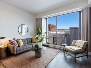 A modern living room with a grey sofa, a round coffee table, a chair, a mirror, and a large window with a city view. - Loft | Quiet Apartment Old Montreal, Courtyard, Full Kitchen (Montréal)