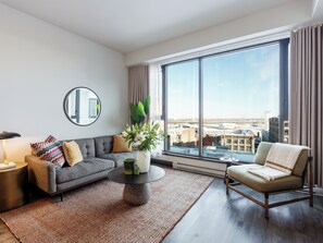 A modern living room with a grey sofa, a round coffee table, and a view of the city through large windows. - Loft | Serviced Apartment Old Montreal, King Bed, Full Kitchen (Montréal)