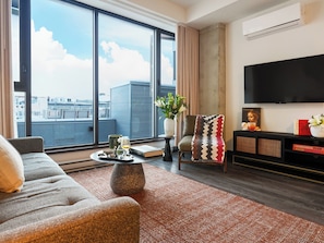 Un salon moderne avec un canapé, une table basse, un téléviseur et une vue sur le paysage urbain grâce à de grandes fenêtres.