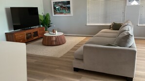 Ein Wohnzimmer mit einer Couch, einem Fernseher auf einer Holzkommode, einem runden Holztisch, einer Pflanze und einem Gemälde an der Wand.