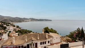 Eine Küstenstadt mit Häusern mit Terrakotta-Dächern, freier Sicht auf das Meer und einer hügeligen Landschaft im Hintergrund.
