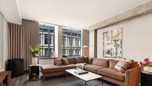 A modern living room with a brown sofa, a coffee table, a large window with curtains, a painting on the wall, and a suitcase on the floor. - One-Bedroom Suite | Bright Apartment Old Montreal, Street View, Open Living (Montréal)