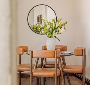Een eetgedeelte met een ronde tafel, vier bruine leren stoelen, een witte vaas met bloemen en een ronde spiegel aan de muur.