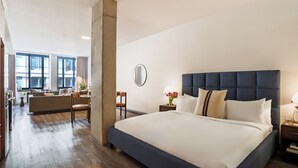 Una habitación de hotel con una cama grande, una zona de estar con sofá y sillones, una mesa de comedor y vistas de la ciudad a través de amplios ventanales.
