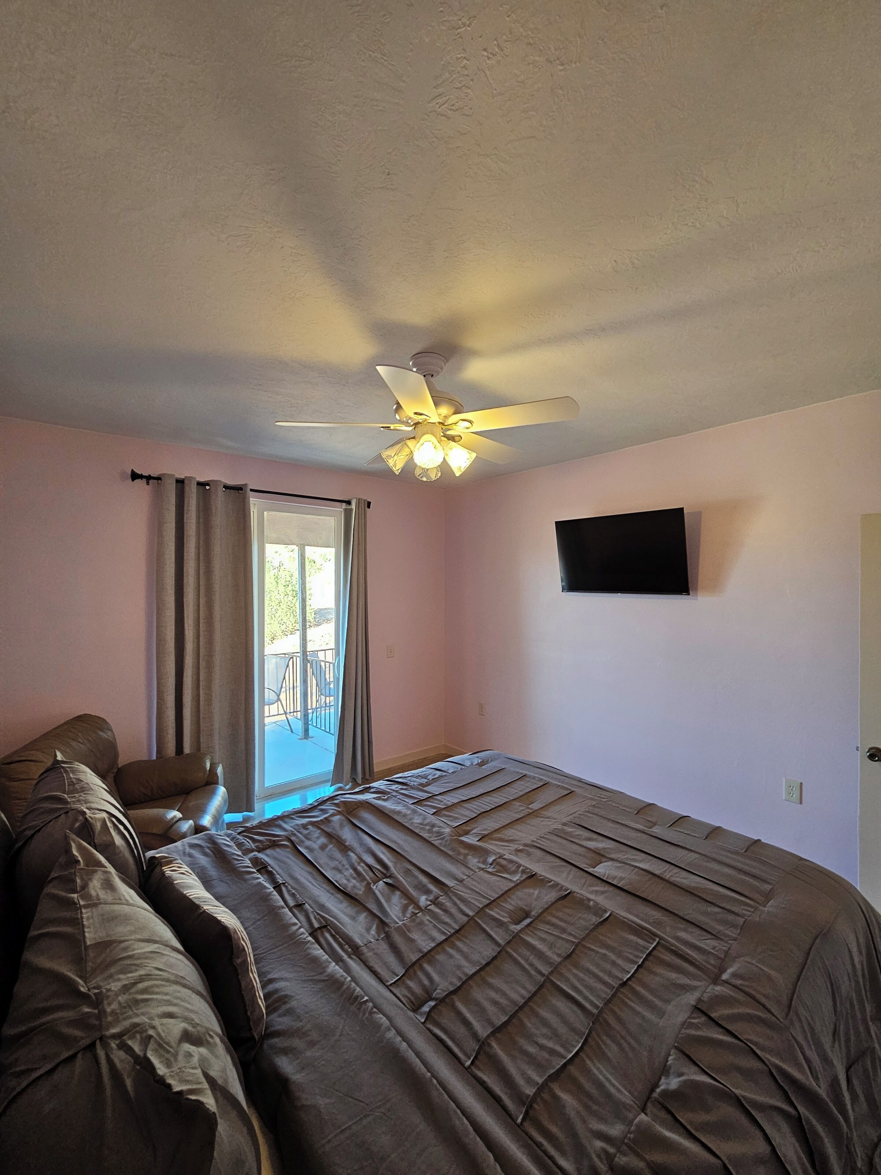 A bedroom with a bed, a ceiling fan, a TV on the wall, and a window with curtains.
