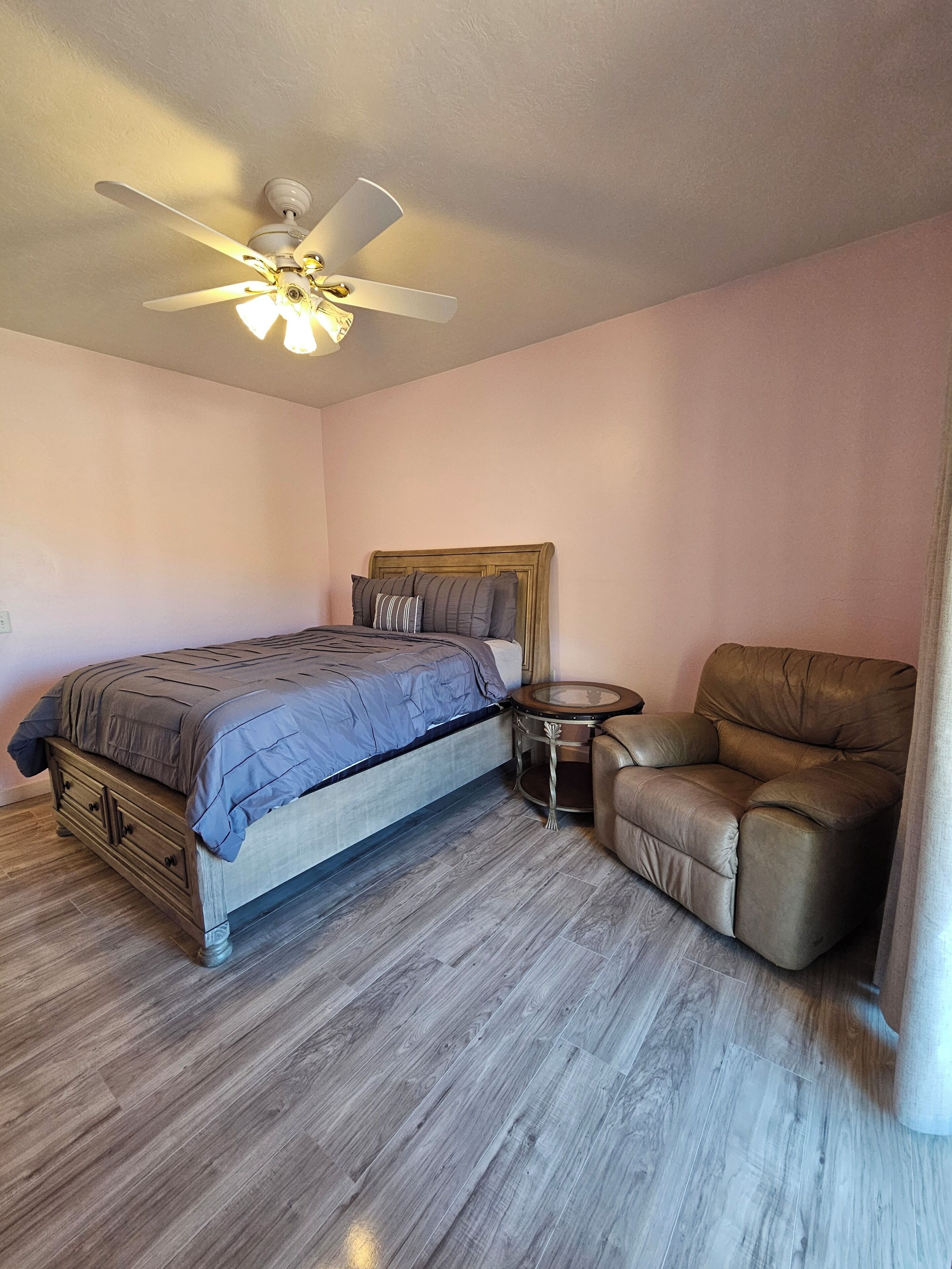 A bedroom with a bed, a chair, a small round table, and a ceiling fan.