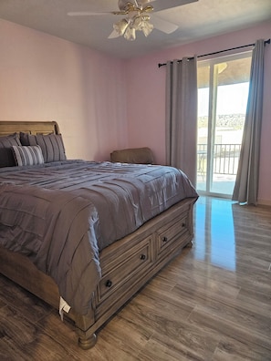 A bedroom with a bed, a wooden dresser, a ceiling fan, and a view through a window.