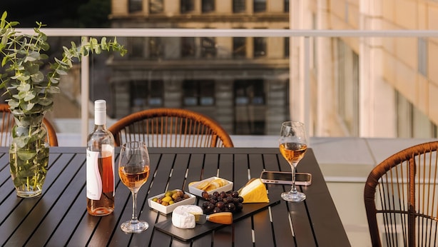 A table with a bottle of wine, glasses, a cheese board, and a vase of greenery on a balcony overlooking a cityscape. - Penthouse | Luxury Residence Old Montreal, Private Terrace, Two Levels (Montréal)