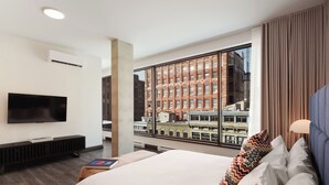 A hotel room with a large bed, a flat-screen TV mounted on the wall, a window with a city view, and a modern lamp. - Penthouse | Luxury Residence Old Montreal, Private Terrace, Two Levels (Montréal)