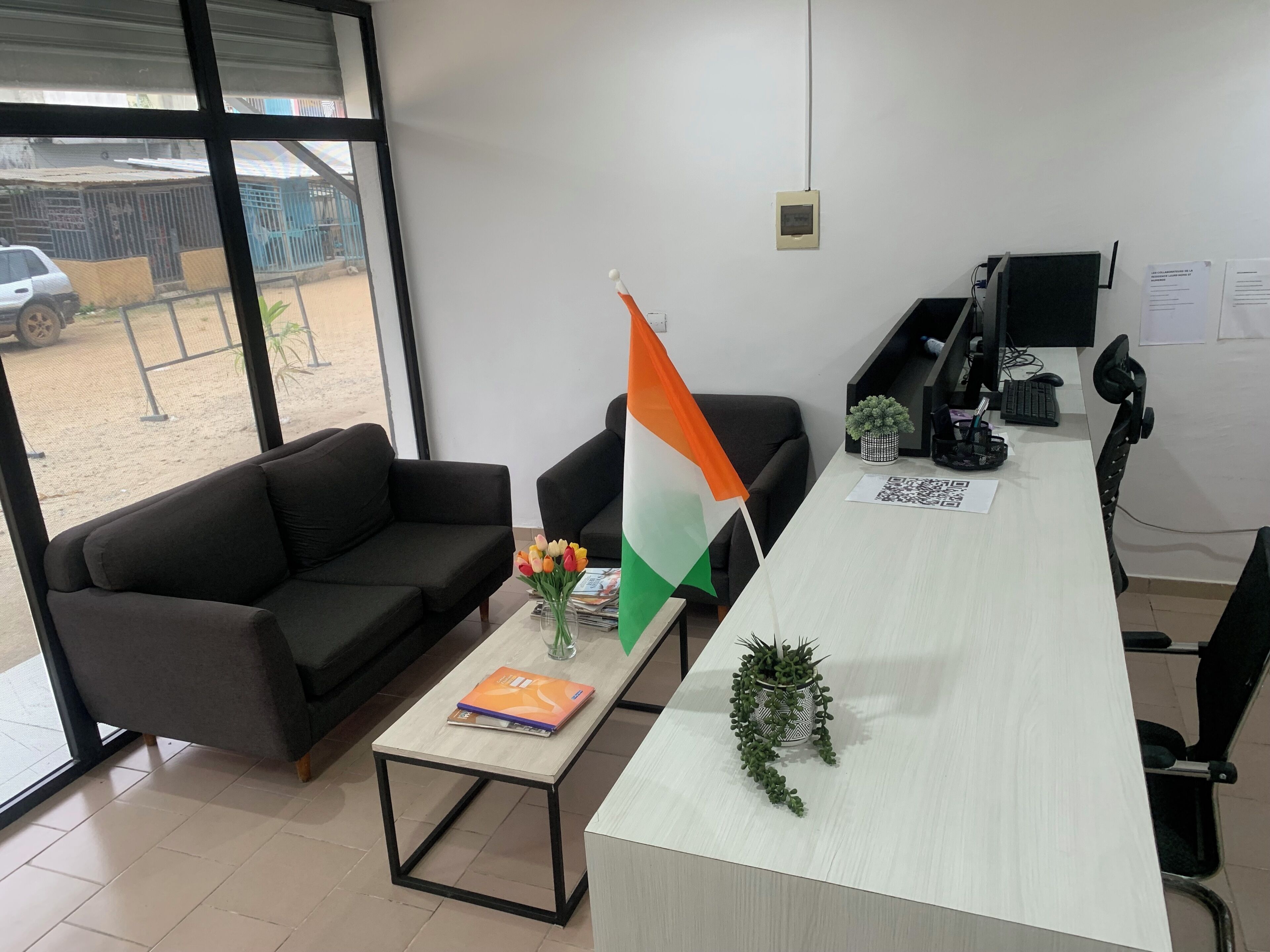 Een kantoor met een lange tafel, twee zwarte banken, een vlag, een kleine plant en een raam met uitzicht op een gebouw en een auto.