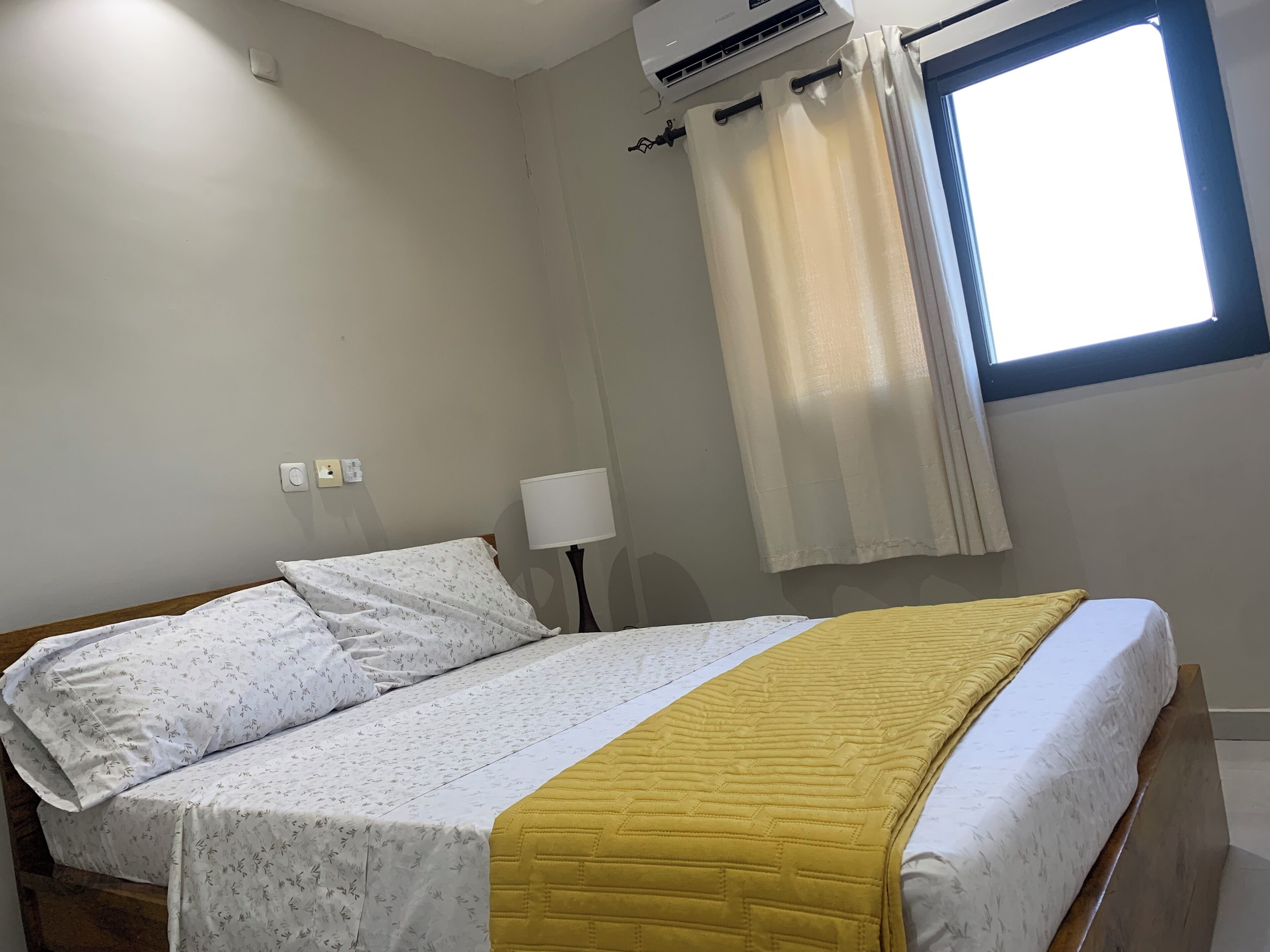 A single bed with a patterned bedspread and pillows, a bedside lamp, an air conditioning unit, a window with curtains, and a wall socket.