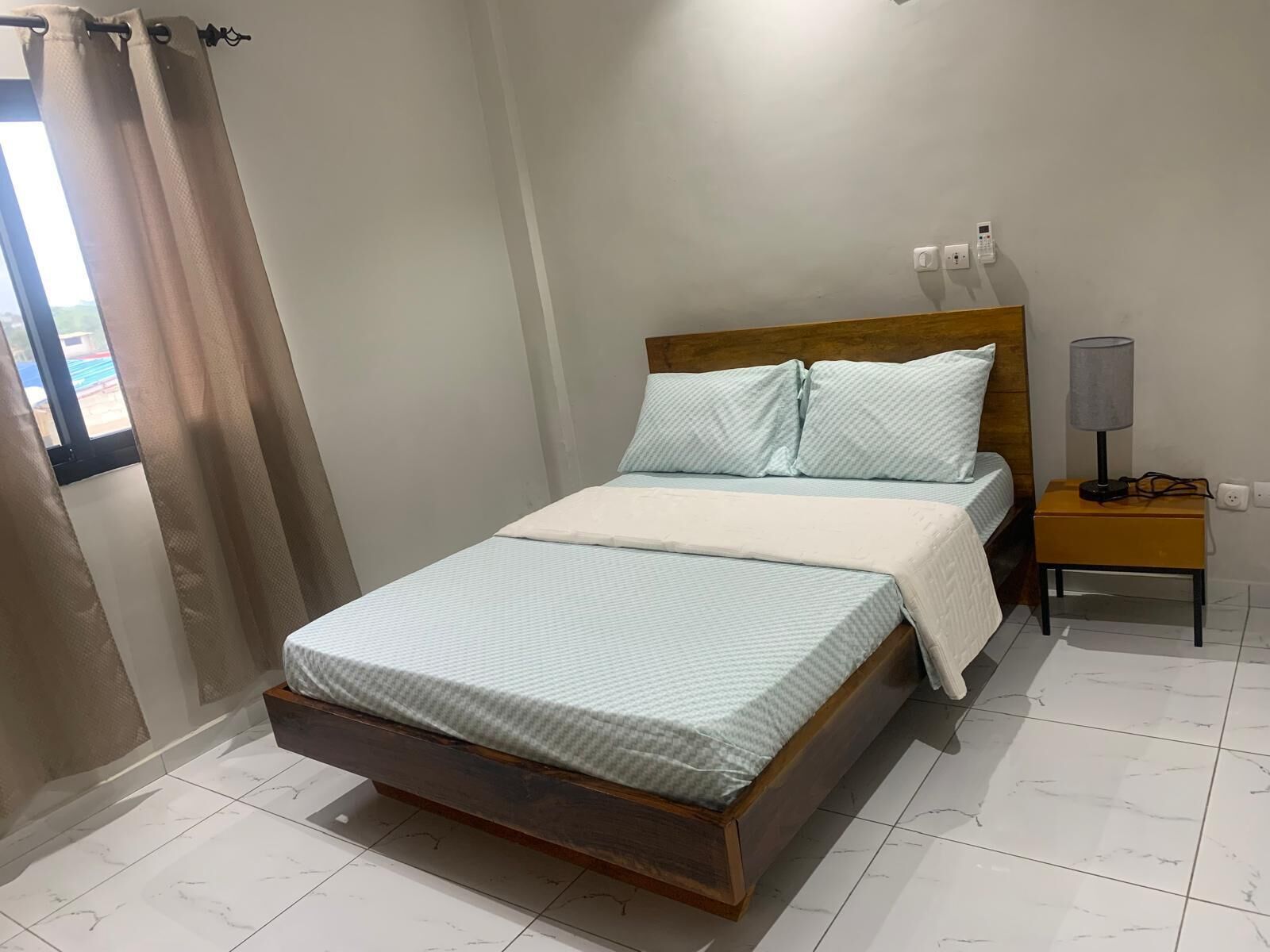A bedroom with a wooden bed, a bedside table with a lamp, and a window with curtains.