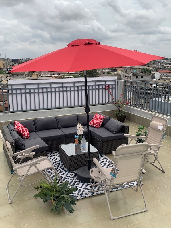 A rooftop terrace with a black sectional sofa, two folding chairs, a red umbrella, and a potted plant.