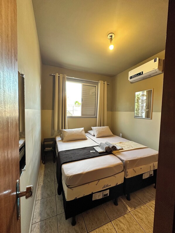 A hotel room with two beds, a window with curtains, an air conditioning unit, and a small table.