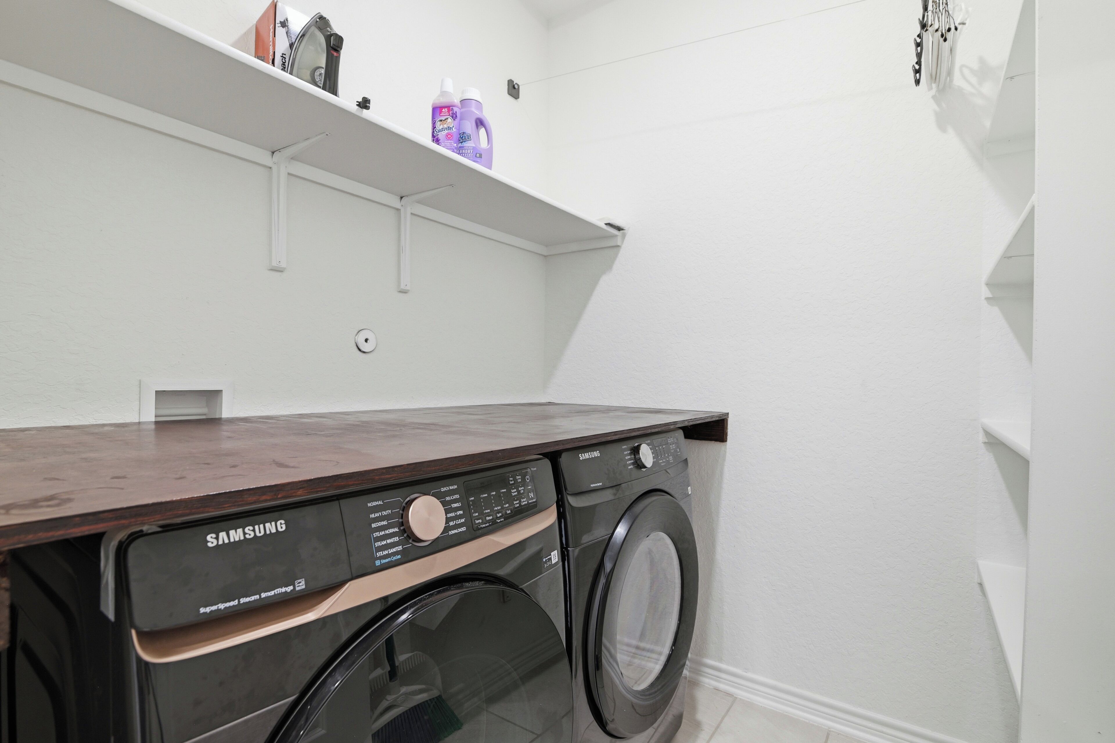 A laundry room with a Samsung SuperSpeed Steam Washer and dryer, a white wall, and a shelf with cleaning supplies.