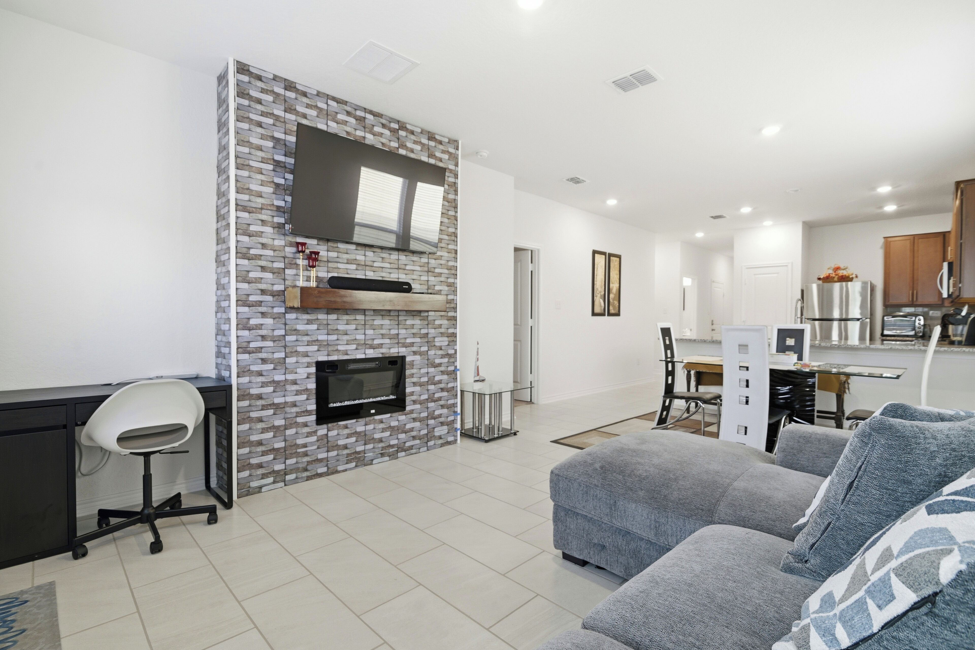 A modern living room with a grey sofa, a TV mounted on a brick wall, a fireplace, and a kitchen area with cabinets and appliances.
