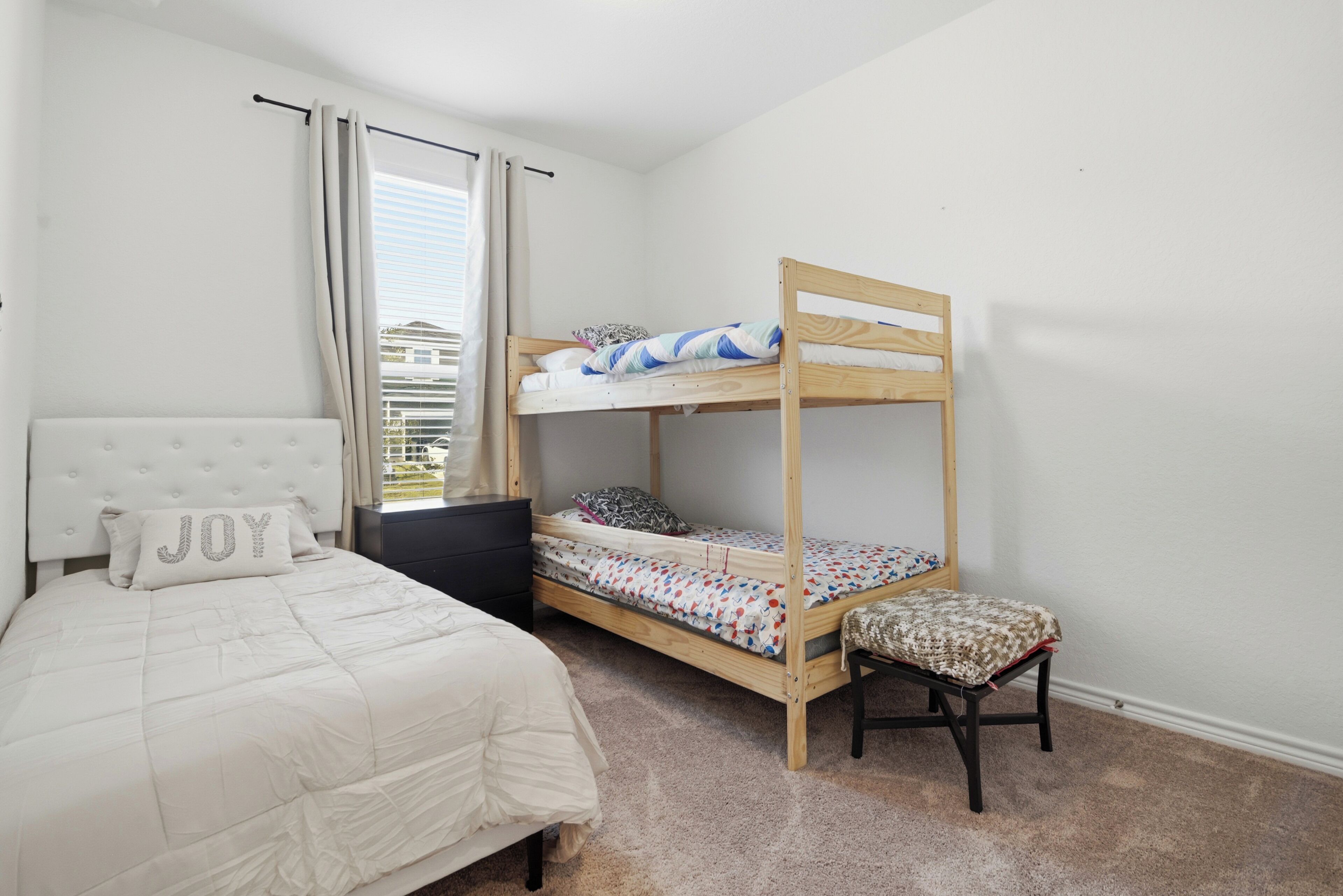 A bedroom with a bunk bed, a single bed, a chair, a nightstand, and a window with curtains.