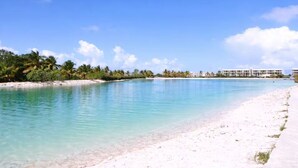 A beach with clear turquoise water, white sand, and a row of white buildings in the background. - 1BR on P. Lake Vista Lake – Concierge S., Pool & Gym (Punta Cana)