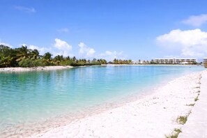 Ein Strand mit klarem türkisblauem Wasser, weißem Sand und einer Reihe weißer Gebäude im Hintergrund.