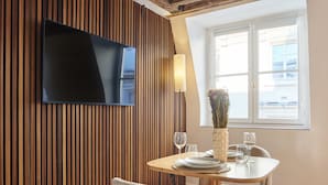 Een eethoek met een wand van houten panelen, een flatscreen televisie, een vaas met bloemen en een raam met zonwering.