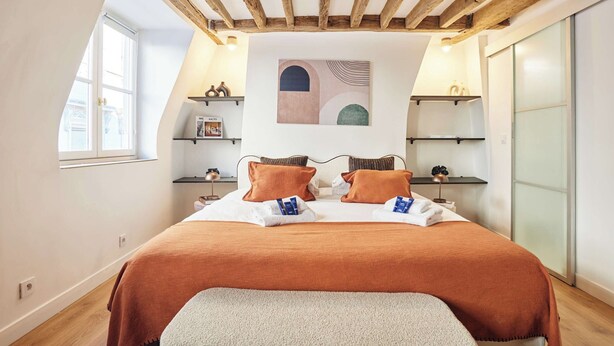 Een slaapkamer met een schuine kap, een bed met oranje beddengoed, een nachtkastje met een lamp, en een wandplank met decoratieve objecten.