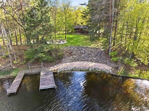 Eine Holzstele führt zu einer Steinbrücke über ruhiges Wasser, umgeben von üppiger Vegetation und einer kleinen Holzhütte im Hintergrund.