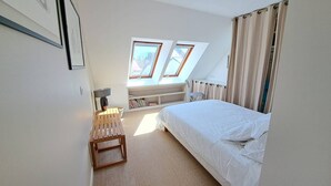 Et soveværelse med en seng, et lille bord, en lampe, en reol med bøger og to ovenlys.