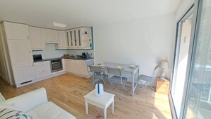 Et kompakt køkken med moderne hårde hvidevarer, et spiseområde med bord og stole, samt en sofa i en lys stue med et stort vindue.