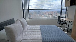 Ett hotellrum med en säng, ett skrivbord och utsikt över stadsbilden genom stora fönster.