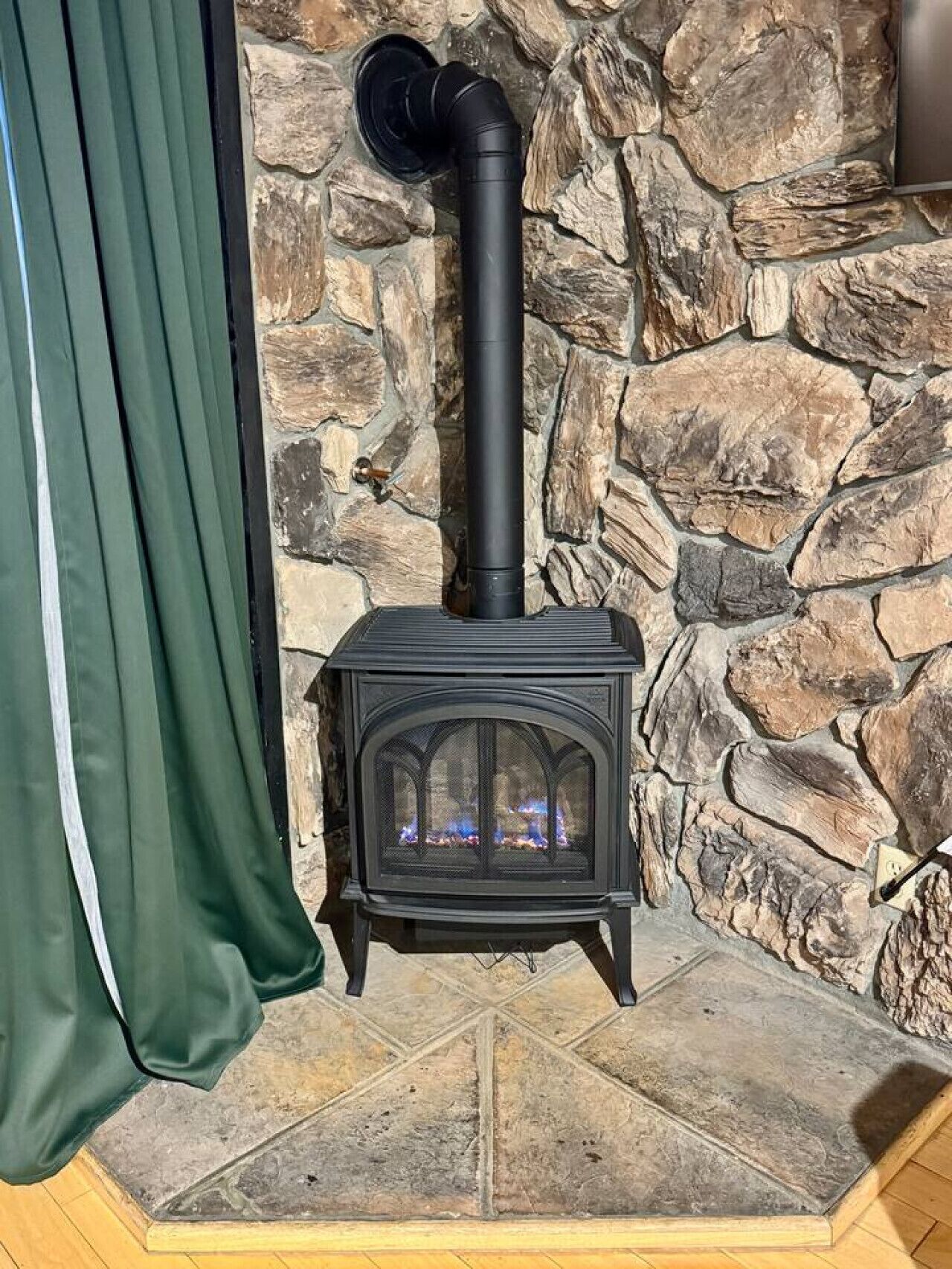 En vedeldad kamin med skorsten, placerad framför en stenmur, och en grön gardin syns delvis.