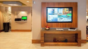 Eine Hotelhalle mit einem großen Flachbildfernseher an einer Holzkonsole, einem Empfangstisch mit Computer und einer Zierpflanze.