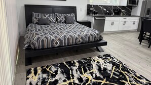 A modern bedroom with a bed, a black and white patterned bedspread, a black and gold rug, a kitchen with white cabinets, and a black and white artwork on the wall. - Modern Luxury Studio Cozy Retreat (Zephyrhills)