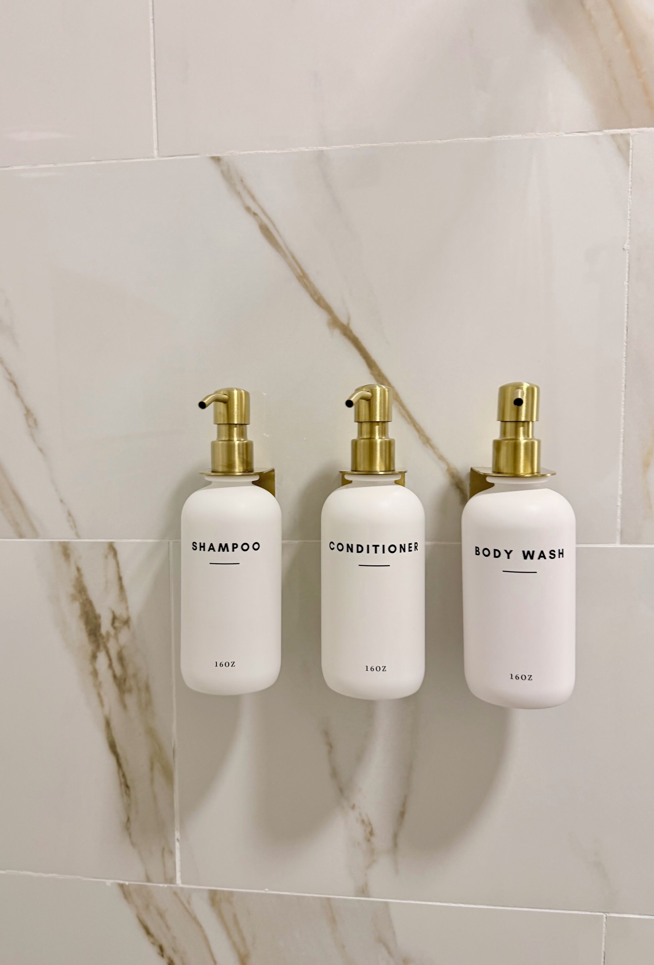 Three white bottles with gold pumps labeled 'SHAMPOO,' 'CONDITIONER,' and 'BODY WASH' on a tiled wall.