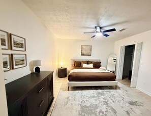 Un dormitorio con cama, mesitas de noche, ventilador de techo, cuadros enmarcados en la pared y puerta a otra habitación.