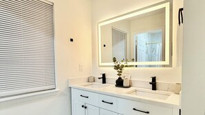 Ein modernes Badezimmer mit einem weißen Waschtisch, einem großen Spiegel und einem Fenster mit Jalousien.