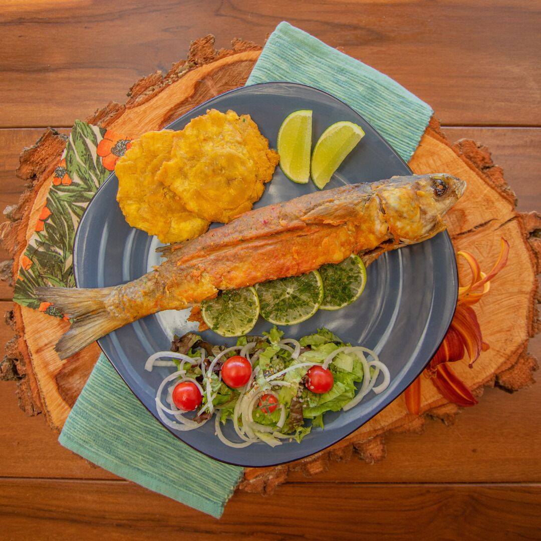 A plate with a whole fried fish, yellow plantain, and a salad with tomatoes and onions.