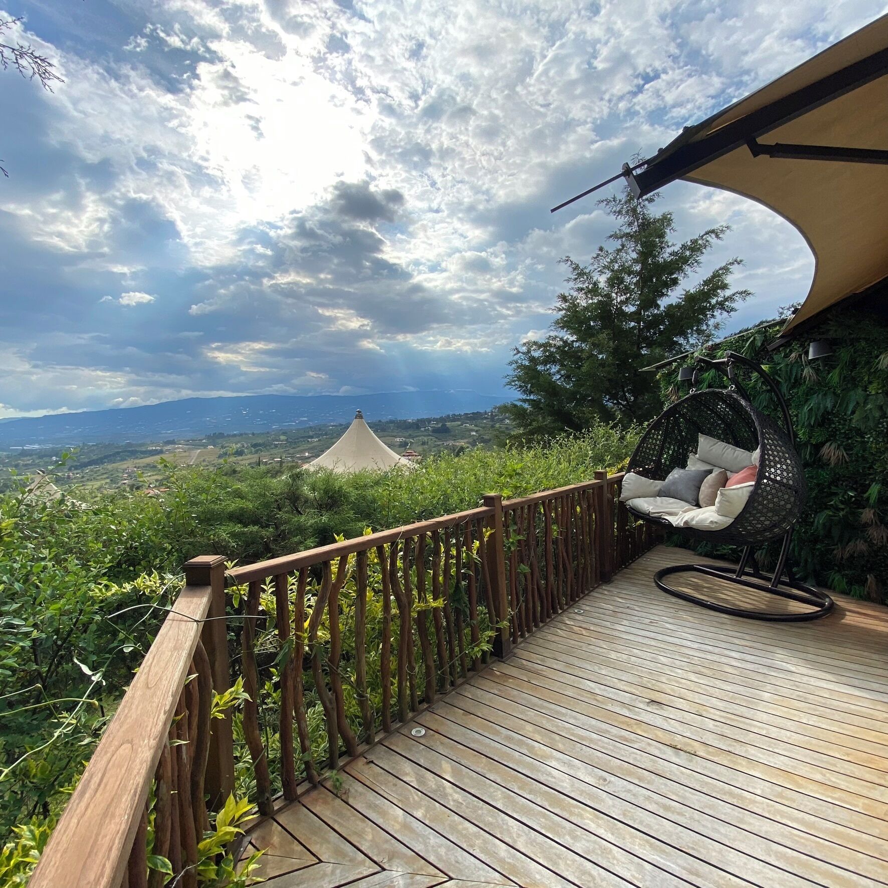 Una terraza de madera con una silla mecedora, vista a un paisaje lejano y una estructura similar a una tienda de campaña.