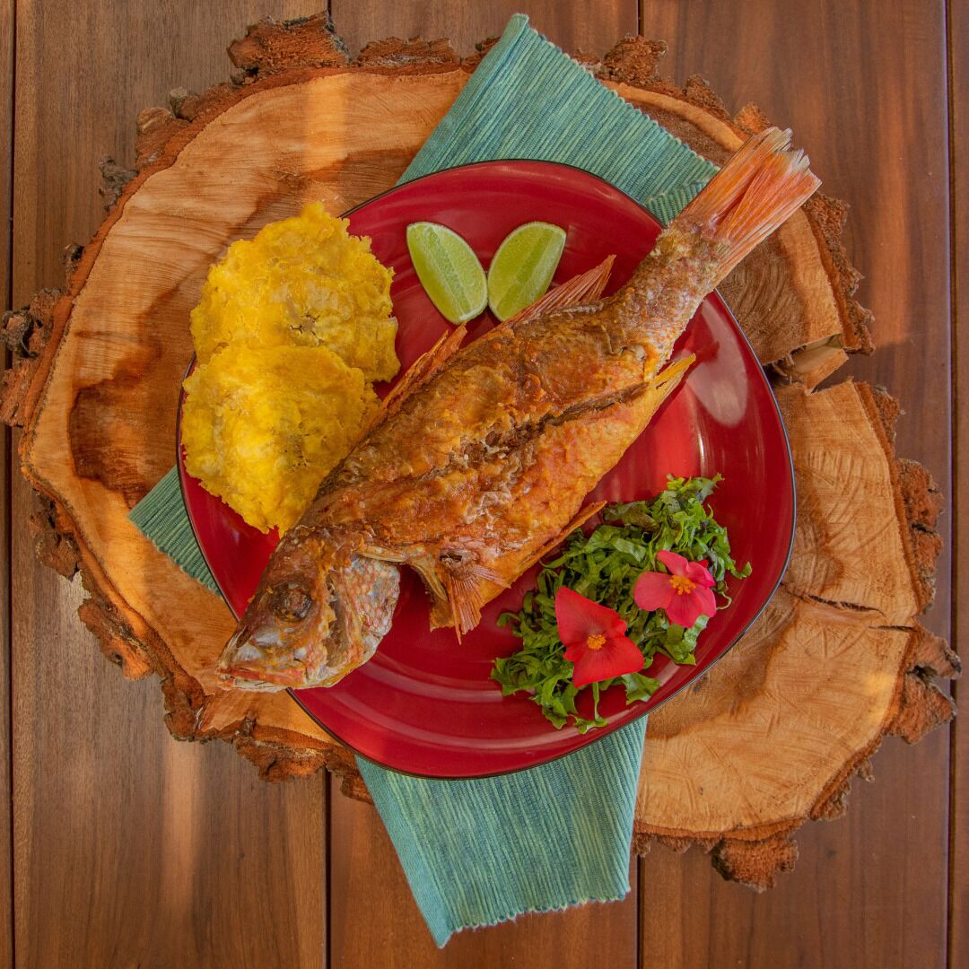 Une assiette contenant du poisson frit, des bananes plantains jaunes et une fleur rouge en guise de décoration, le tout posé sur une table en bois.